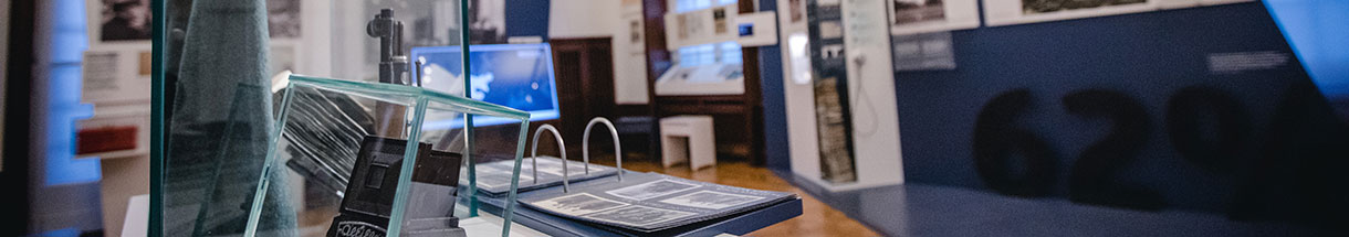 Blik in een tentoonstellingsruimte met veel informatieborden aan de wanden, op de tafel een open map met foto's