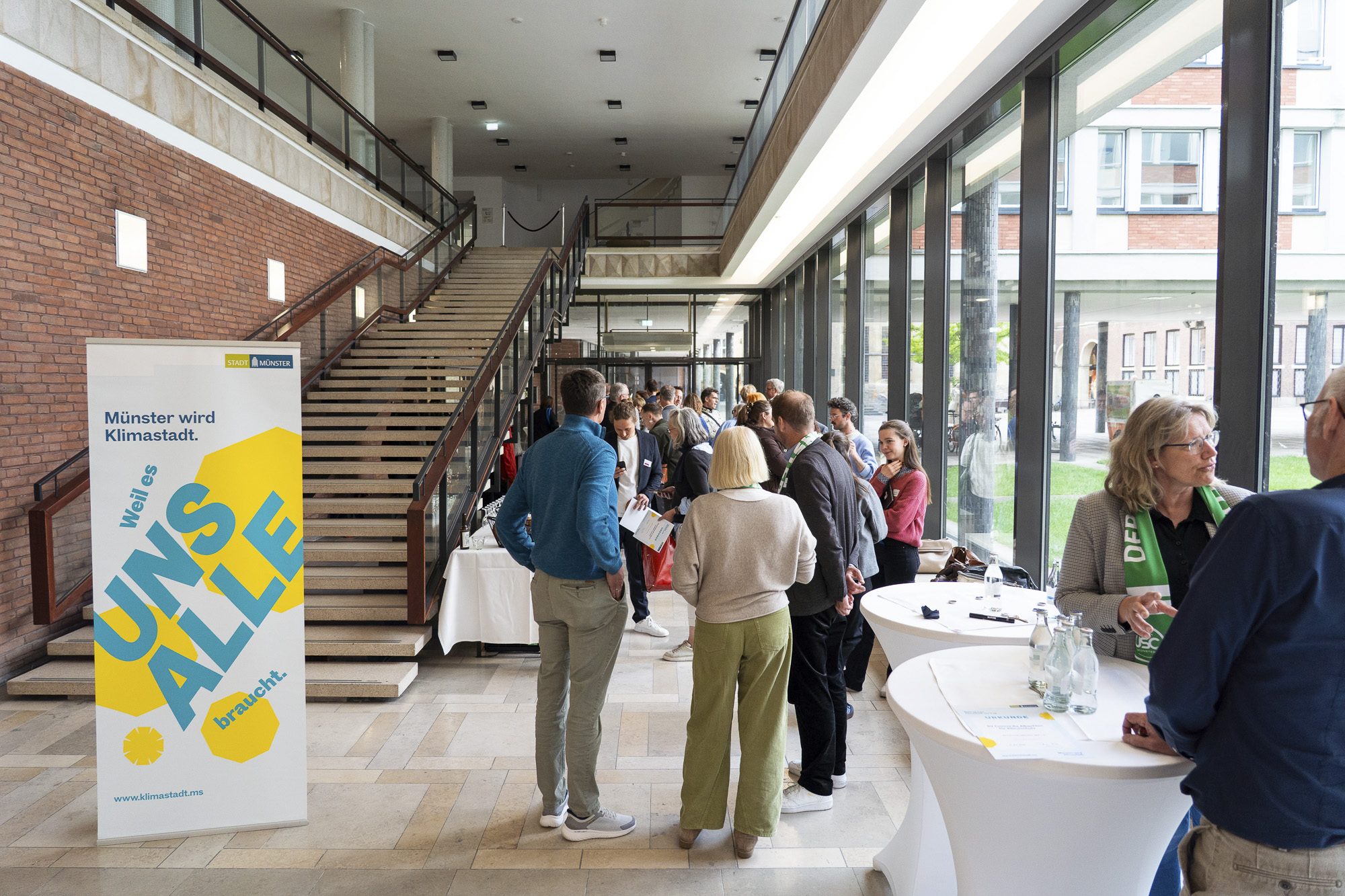 Menschen stehen in einem hellen Foyer mit großen Fenstern an Stehtischen und einem Informationsbanner mit der Aufschrift 'Münster wird Klimastadt'