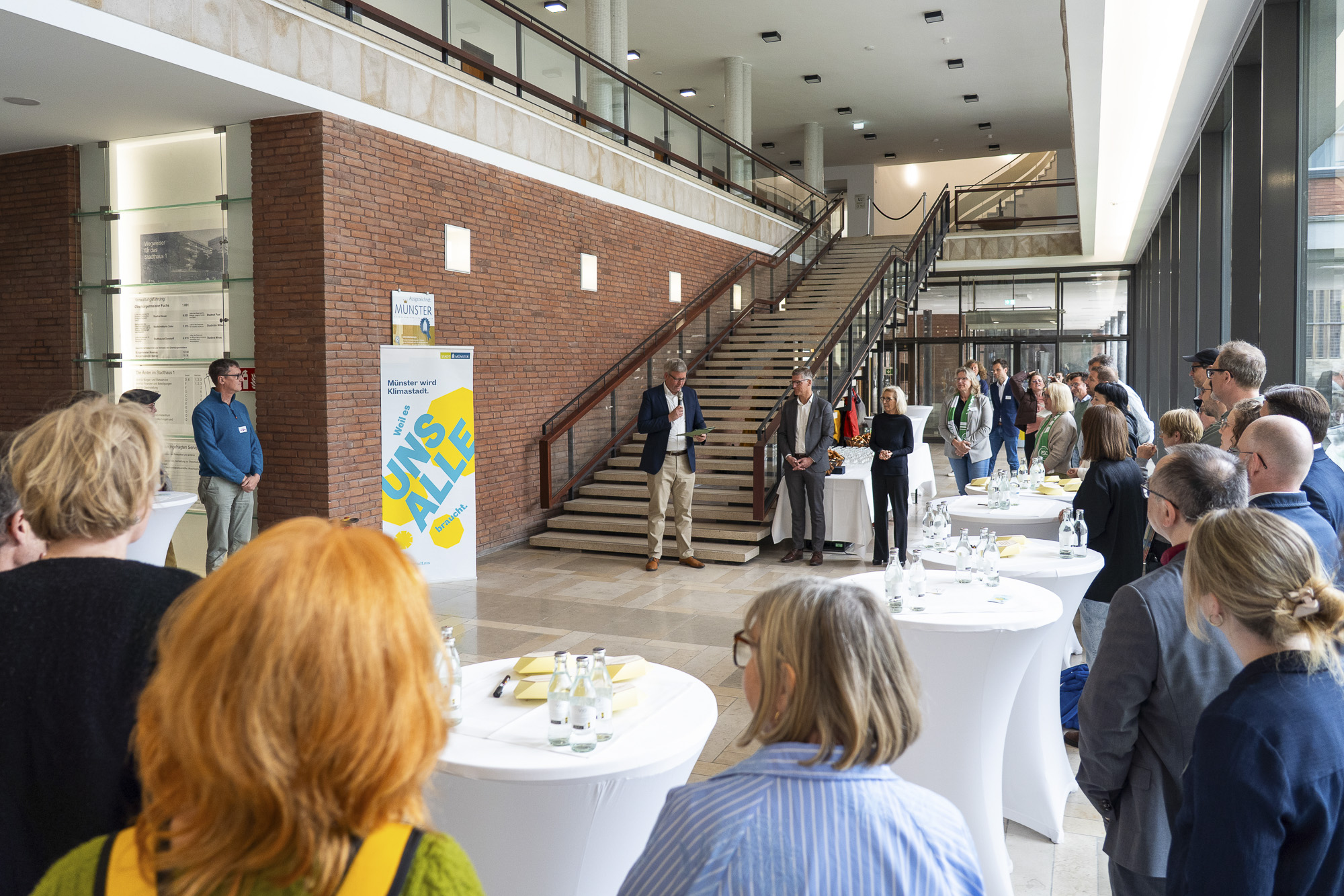 Menschen stehen in einem Foyer mit Treppe und hören einem Mann zu, der vor einem Banner spricht