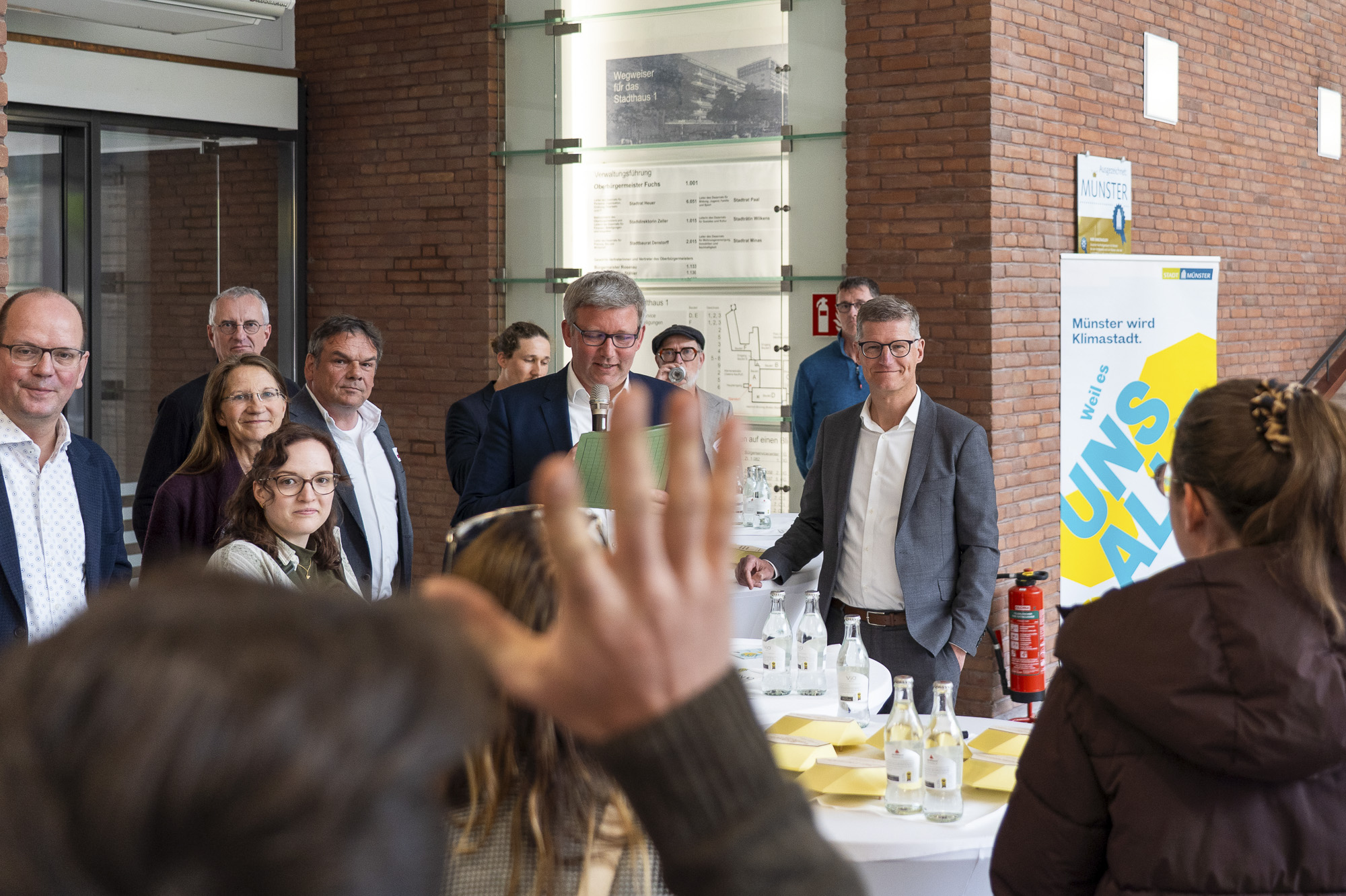 Gruppe von Menschen steht in einem Raum mit Backsteinwänden, eine Person im Vordergrund hebt die Hand, auf einem Tisch stehen Wasserflaschen und Gläser, im Hintergrund ein Plakat mit dem Text 'Münster wird Klimastadt'