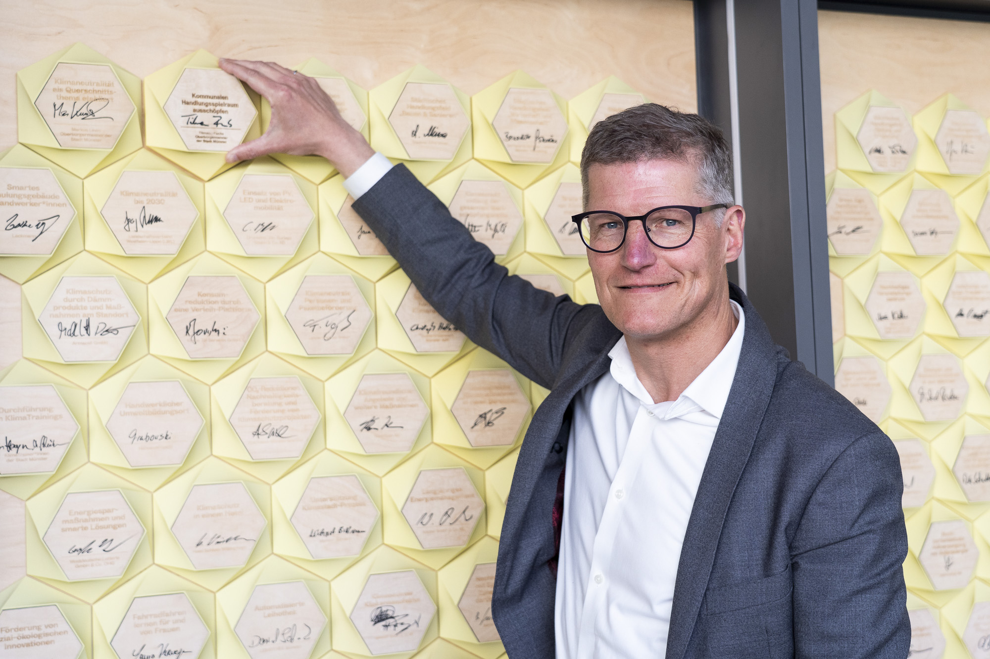 Mann in Sakko und weißem Hemd steht vor Wand mit vielen sechseckigen Plaketten, die Unterschriften tragen, und berührt eine Plakette