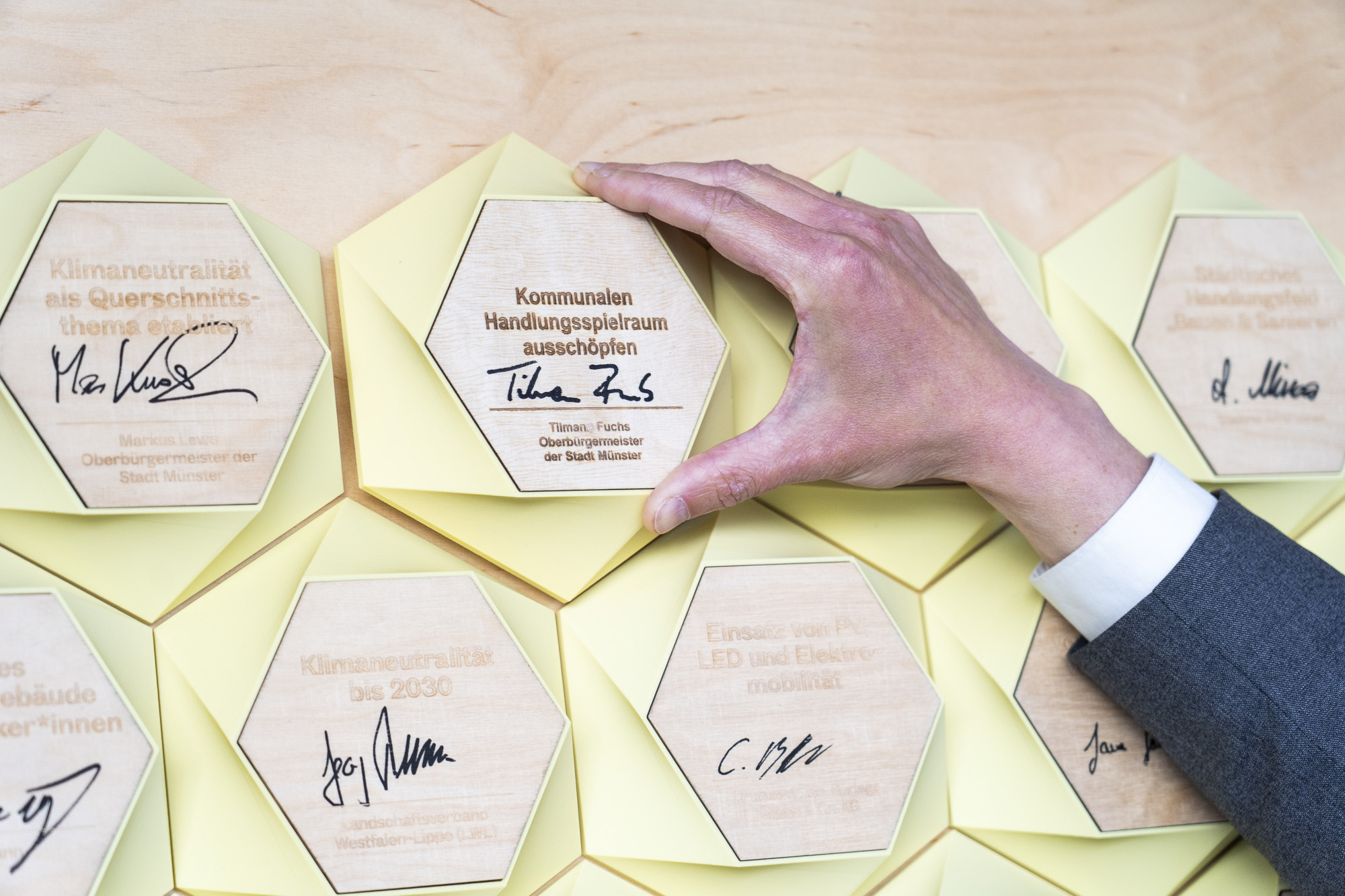 Hand hält sechseckige Holztafel mit Text und Unterschrift über weitere sechseckige Holztafeln auf hellem Untergrund