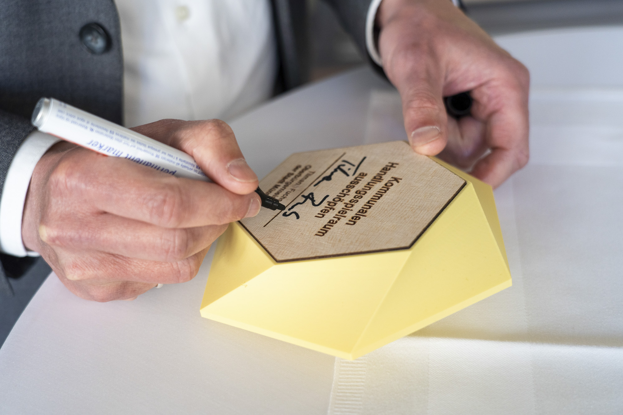 Hände eines Mannes in Anzug, der mit einem schwarzen Stift auf einem sechseckigen, gelbe Kachel schreibt