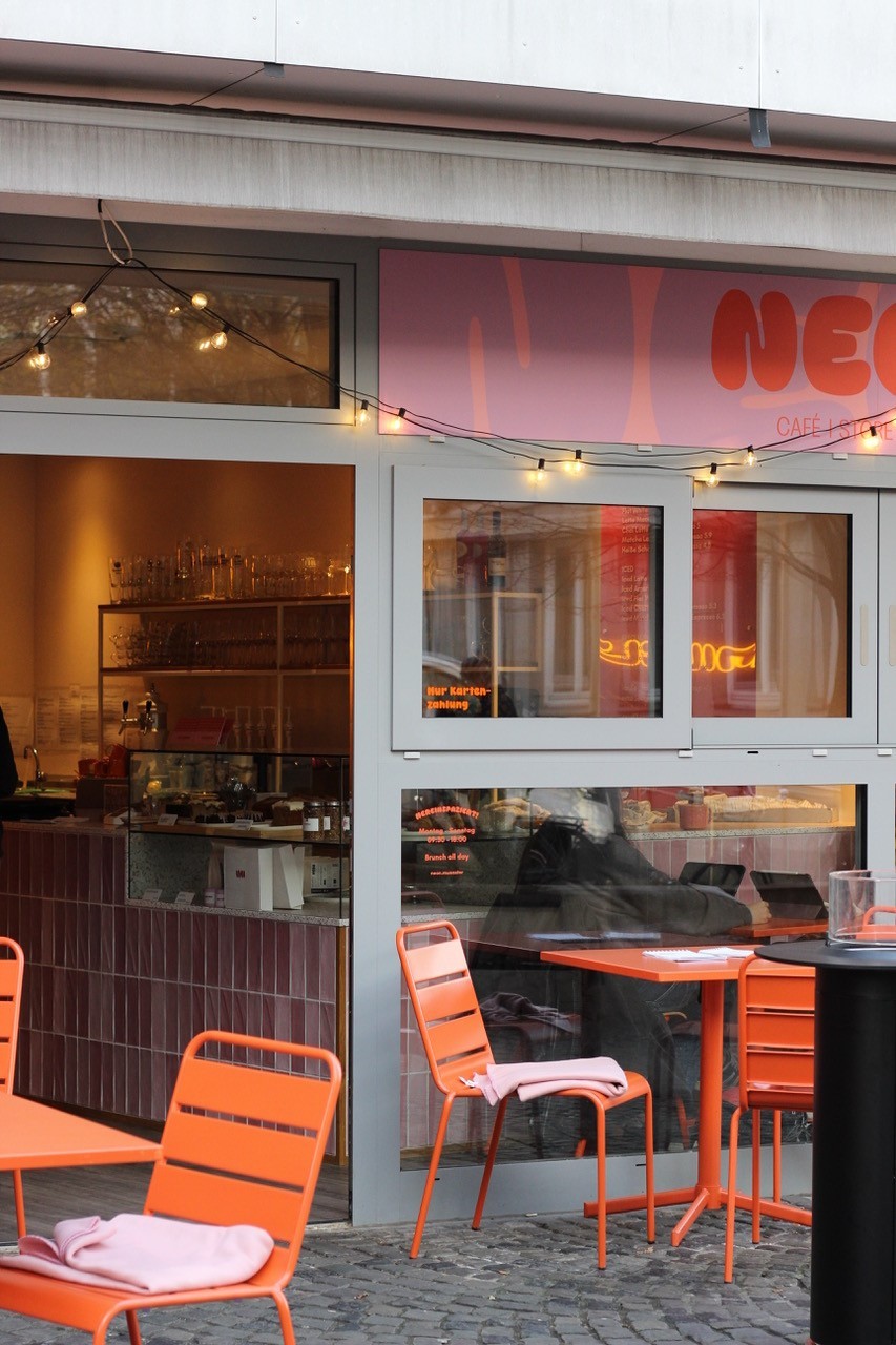 Ein Foto von einem Café vor dem orangene Tische und Stühle stehen