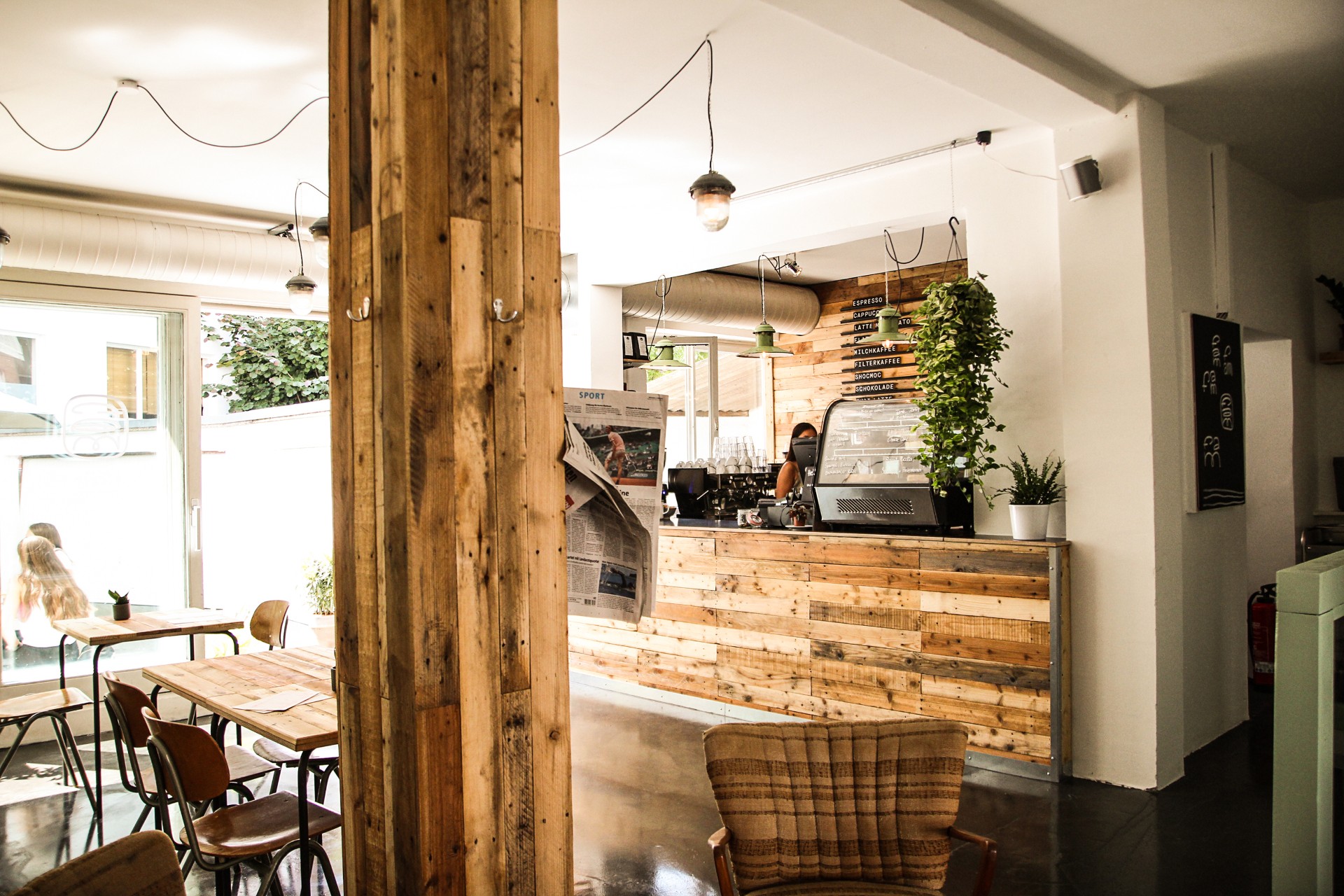 Innenansicht einer Café-Bar. Mit Holztischen und einem Holzbalken in der Raummitte. Hinter der Bar steht eine Frau.