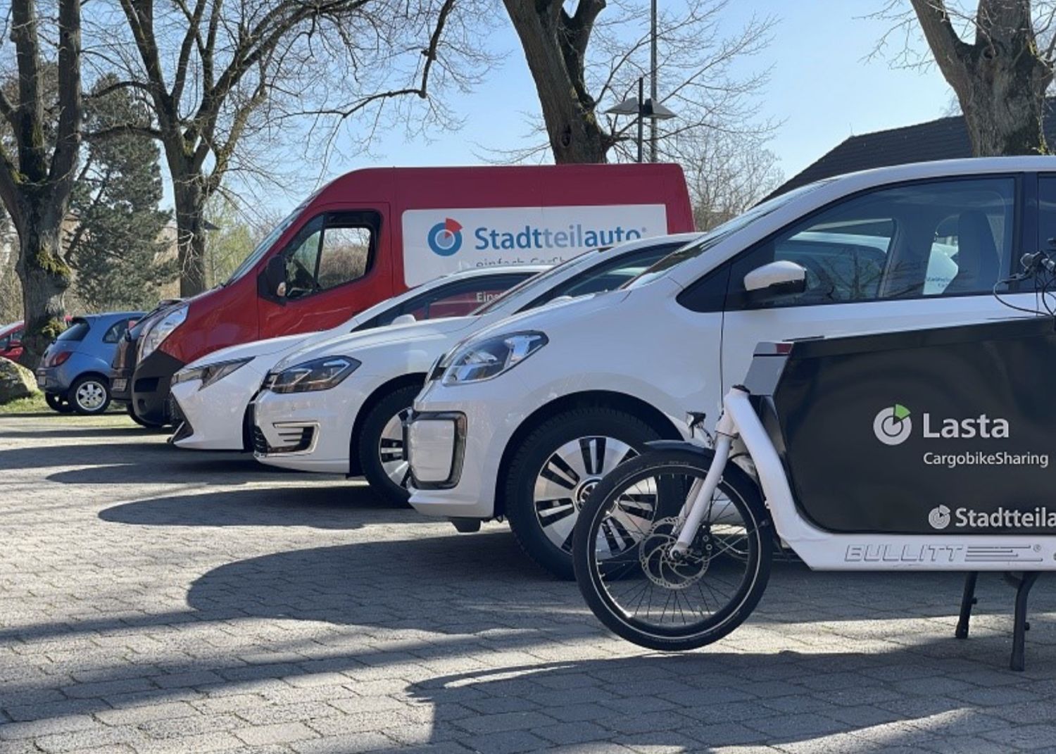 Mehrere Fahrzeuge einer Carsharing-Flotte, darunter ein weißes Lastenfahrrad mit "Lasta CargobikeSharing"-Beschriftung und ein roter Transporter mit "Stadtteilauto"-Logo, stehen nebeneinander auf einem Parkplatz.