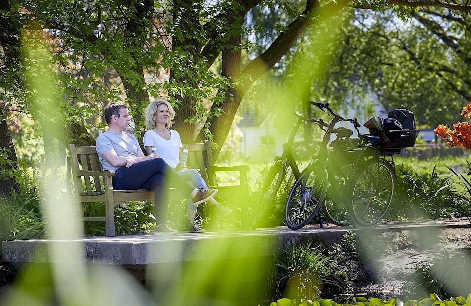Eine Mann und eine Frau sitzen auf einer Parkbank vor einem Teich. Neben der Bank steht ein Fahrrad.