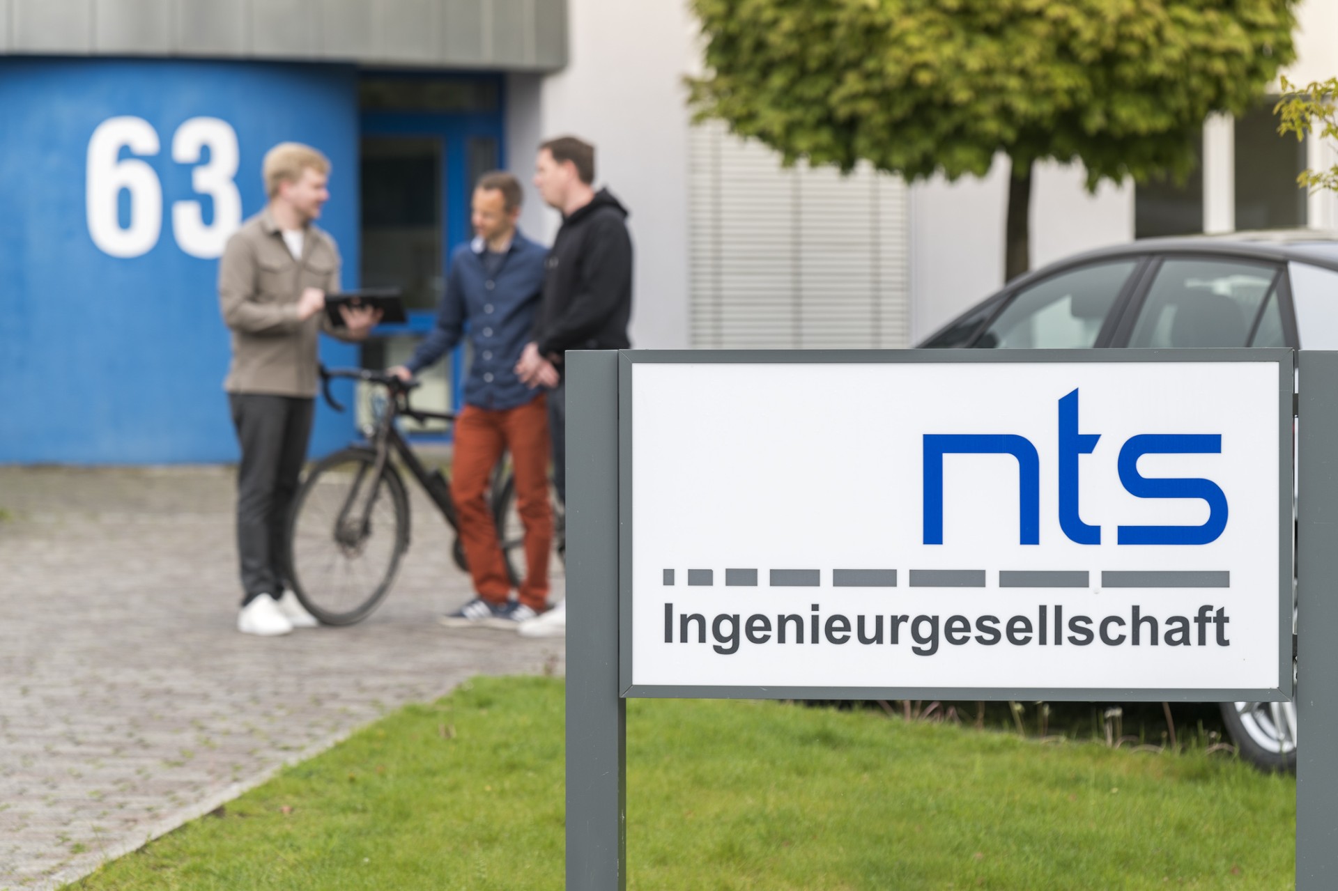 Ein Foto einer Einfahrt mit dem Schild "nts Ingenieurgesellschaft", im Hintergrund drei Männer und ein Gebäude