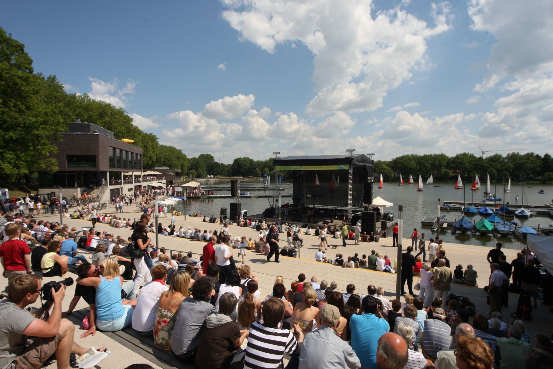 Menschen sitzen auf Stufen am Ufer eines Sees vor einer Bühne mit Segelbooten im Wasser und bewölktem Himmel
