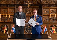 Onno van Veldhuizen und Markus Lewe stehen im Friedenssaal und halten die Partnerschaftsurkunden in den Händen, vor ihnen auf dem Tisch stehen rechts und links jeweils die deutsche und die niederländische Flagge.