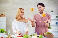 Mit Spaß selber kochen und Mahlzeiten zubereiten. Frau und Mann kochen gemeinsam. Mann wirft Orangen hoch, Frau schneidet Gemüse.